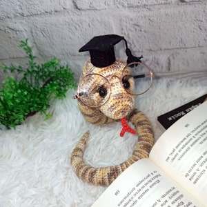 Puede incluir: Una serpiente de crochet con un gorro de graduación negro y gafas redondas. La serpiente es marrón y beige con una lengua roja. La serpiente está sentada en un libro con texto en un idioma extranjero.