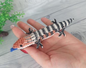 May include: A handmade crochet lizard in a realistic style. The lizard features a striped black and white body, an orange head, and a blue tongue. The lizard is lying on a hand.