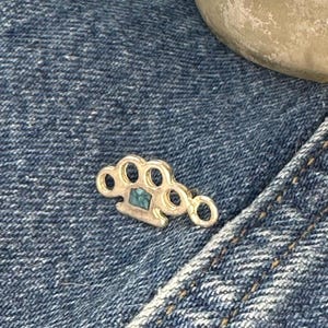 May include: A silver-colored brass knuckle pin with a square, teal-colored stone set in the center. The pin is resting on a blue denim fabric.