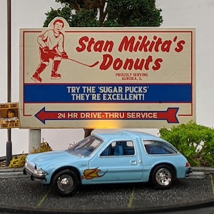 May include: A detailed miniature diorama showcasing a light blue toy car with a flame decal, positioned before a vintage "Stan Mikita's Donuts" sign. The sign features text and a hockey player illustration. A street lamp and small tree are also included.