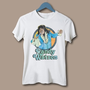 Puede incluir: Camiseta blanca con una ilustración vintage de Randy Watson con un traje azul y un micrófono. El gráfico está dentro de un diseño circular con detalles en naranja y amarillo, y el nombre "Randy Watson" impreso debajo en una fuente estilizada.