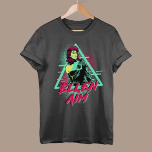 May include: A dark gray t-shirt with a retro-style graphic of a woman with red hair in a black leather jacket. The graphic is set in a turquoise triangle with the text "ELLEN AIM" in pink and red letters.