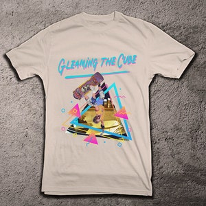 Op de afbeelding: Een wit T-shirt met een grafische print van een persoon die skateboardt in een driehoek met de tekst "GLEAMING THE CUBE" in blauw en roze.