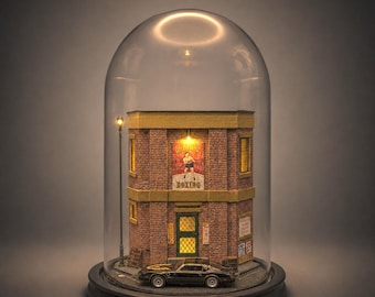 Puede incluir: Un diorama de edificio de ladrillo en miniatura bajo una cúpula de cristal, con un letrero de gimnasio de boxeo, ventanas iluminadas y un coche deportivo negro aparcado delante. La estructura tiene bordes amarillos, una puerta verde y una farola.