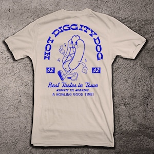 May include: A white t-shirt with a blue graphic of a hot dog character with the text "Hot Diggity Dog" in a circle around it. The text "Best Tastes in Town" is below the character, with the text "Midnite Til Morning" and "A Howling Good Time!" below that.