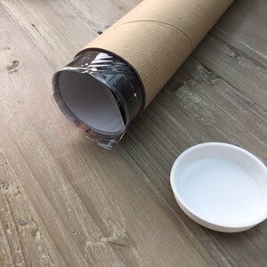 May include: A rolled-up poster or print is being shipped in a brown cardboard tube. The tube is lying on a wooden surface. A white plastic lid is also visible.