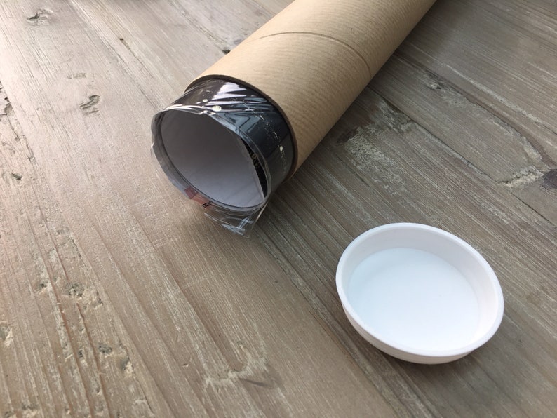 May include: A rolled-up poster or print is being shipped in a brown cardboard tube. The tube is lying on a wooden surface. A white plastic lid is also visible.