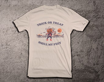 May include: A white t-shirt with a graphic of a pumpkin wearing a cowboy hat and holding a gun. The text "Trick or Treat" is above the graphic and "Smell My Feet" is below the graphic.