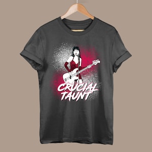 Puede incluir: Una camiseta gris oscuro con un estampado gráfico de una mujer tocando un bajo. El gráfico es en blanco y negro con acentos rojos. El texto "CRUCIAL TAUNT" está impreso en rojo y blanco.