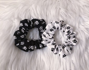 May include: Two scrunchies with paw print patterns. One scrunchie is black with white paw prints and the other is white with black paw prints.