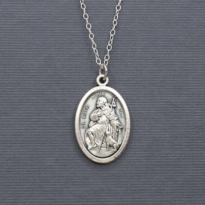 May include: Silver chain necklace with an oval pendant featuring a depiction of Saint Christopher carrying a child. The pendant has the words "St. Christopher" and "Protect Us" inscribed on it.