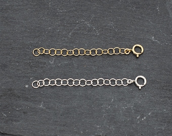 May include: Two chain extenders, one gold and one silver, on a black slate background. Both extenders have a lobster clasp.
