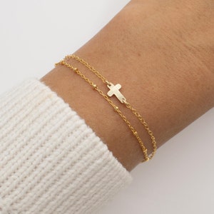 May include: A gold chain bracelet with a small cross charm. The bracelet is layered with two chains, one with small beads and one with a more delicate chain.
