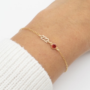 May include: A gold chain bracelet with a red gemstone and the number 2022 in gold lettering.