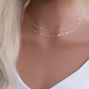 May include: Two silver necklaces layered on a model's neck. The top necklace is a delicate chain, while the bottom necklace features a slightly thicker chain with oval-shaped links.