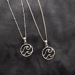 May include: Two silver necklaces with delicate chains and circular pendants featuring a wave design. The necklaces are displayed on a gray fabric background.
