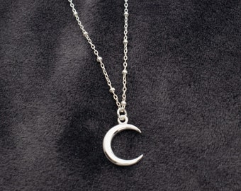 May include: A silver crescent moon pendant necklace on a silver chain with small beads. The necklace is on a dark gray fabric background.