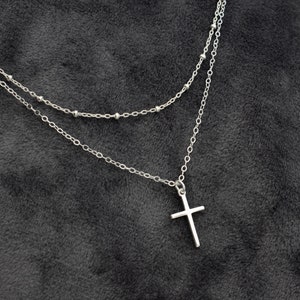 Puede incluir: Un collar de cadena plateado de dos capas con un colgante de cruz plateado.