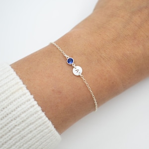 May include: A silver chain bracelet with a small blue gemstone and a silver charm with the letter 'A' engraved on it.