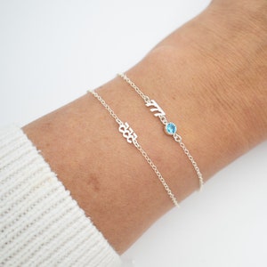 May include: Two silver bracelets with a small blue gemstone on one bracelet. The top bracelet has the word "Mia" on it and the bottom bracelet has the number "333" on it.