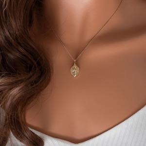 May include: A gold chain necklace with a small, round pendant featuring a detailed design. The pendant is worn on a model with brown hair.