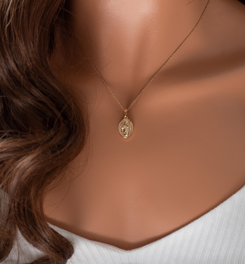 May include: A gold chain necklace with a small, round pendant featuring a detailed design. The pendant is worn on a model with brown hair.
