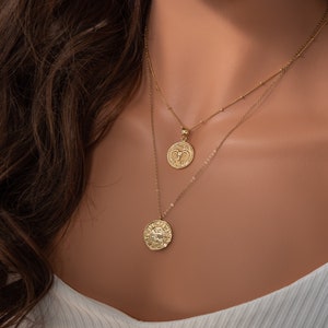 May include: Two gold necklaces with zodiac pendants. The top necklace has a small, round pendant with a ram design. The bottom necklace has a larger, round pendant with a detailed design.