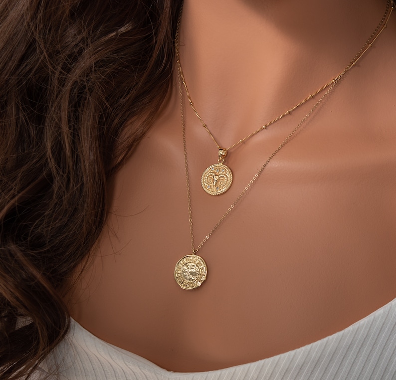May include: Two gold necklaces with zodiac pendants. The top necklace has a small, round pendant with a ram design. The bottom necklace has a larger, round pendant with a detailed design.
