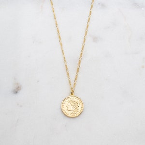 May include: A gold chain necklace with a gold coin pendant featuring a profile of a woman's face. The coin has a raised rim and intricate details.