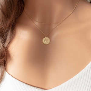 May include: A gold chain necklace with a round coin pendant featuring a woman's profile. The necklace is layered with a delicate chain.