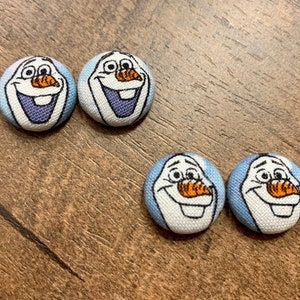 May include: Four round fabric buttons with a blue and white ombre background featuring a cartoon image of Olaf from the movie Frozen. The buttons have a black outline and a white smile.