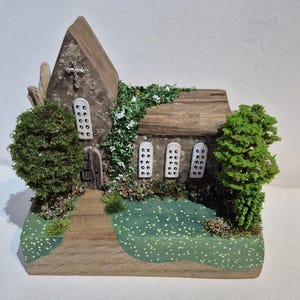 May include: A miniature wooden church model with a stone facade and a wooden shingle roof. The model features a cross, small windows, a wooden door, and miniature trees and greenery. The base is painted green with white flower accents.