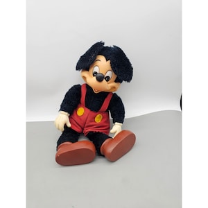 May include: A plush doll featuring a character with black fur, large ears, and a tan face. The doll wears red shorts with yellow buttons and brown shoes. The character has white gloves and a black nose, with a soft, textured body and a moulded face.