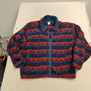 May include: A long-sleeve fleece jacket featuring a repeating horizontal pattern of red, dark blue, and teal stripes. The jacket has a full-length front zipper, a solid teal collar, and two side pockets with zipper closures along the bottom hem.