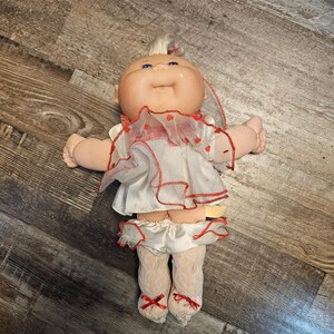 May include: A doll with a round, peach-toned face, blue eyes, and light-colored yarn hair. The doll wears a white sleeveless top with a sheer, ruffled overlay featuring small red hearts and red trim, matching ruffled shorts, and white lace tights.