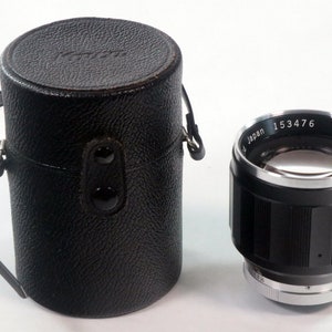 May include: A black Kowa camera lens with the number 153476 and the word "Japan" printed on the silver rim. The lens is next to a black leather case with a handle and two black buttons.