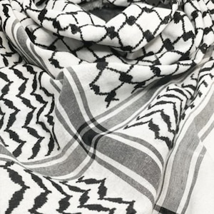 May include: A white and black keffiyeh scarf with a geometric pattern. The scarf features a repeating design of black diamonds and zigzags against a white background. The fabric is folded and draped, showing the texture and detail.