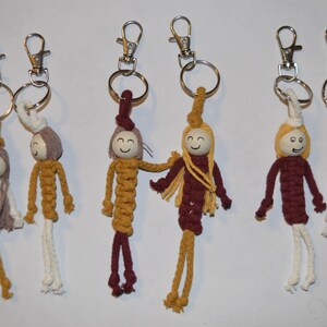 Op de afbeelding: Zes handgemaakte macramé sleutelhangers met kleine houten kralen als hoofdjes en geschilderde lachende gezichtjes. Elke figuur heeft garen haar en een lichaam van geknoopt koord in mosterdgeel, crème en bordeaux, bevestigd aan zilverkleurige karabijnhaken.