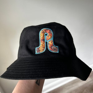 May include: Black bucket hat with a large embroidered letter "A" in orange, yellow, and blue on the front.