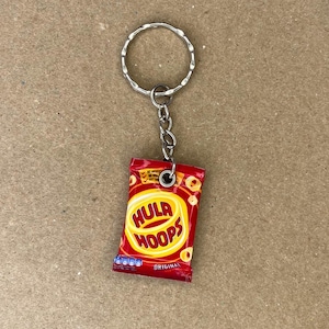 May include: A keychain shaped like a bag of Hula Hoops, a popular brand of snack rings. The bag is red and yellow with the words "Hula Hoops" in white letters. The keychain is attached to a silver key ring.