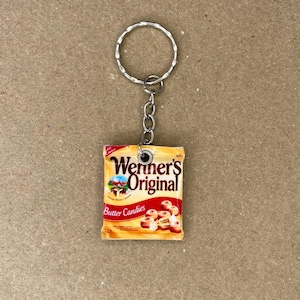 May include: A keychain featuring a miniature replica of a Werner's Original Butter Candies packet. The keychain is made of metal and has a silver finish. The packet is yellow with red and white text that reads "Werner's Original Butter Candies".