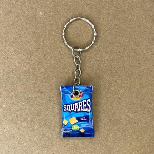 May include: A blue and white bag of Squares Salt & Vinegar potato chips with a silver keychain attached. The bag is a miniature replica of a real bag of chips.