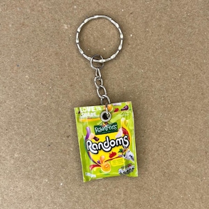 May include: A keychain shaped like a small bag of Rowntree's Randoms sweets. The bag is green and yellow with the words "Rowntree's Randoms" printed on it. The keychain has a silver ring and chain.