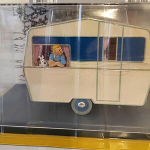 Peut inclure: Une caravane miniature cr&egrave;me avec des accents bleus et marron. Une figurine d'une personne avec un chien est visible dans la fen&ecirc;tre. La caravane a une seule roue et est enferm&eacute;e dans un pr&eacute;sentoir en plastique transparent.
