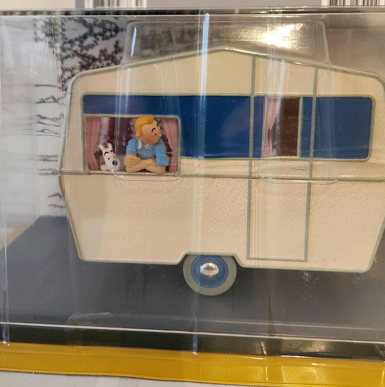 Peut inclure: Une caravane miniature cr&egrave;me avec des accents bleus et marron. Une figurine d'une personne avec un chien est visible dans la fen&ecirc;tre. La caravane a une seule roue et est enferm&eacute;e dans un pr&eacute;sentoir en plastique transparent.
