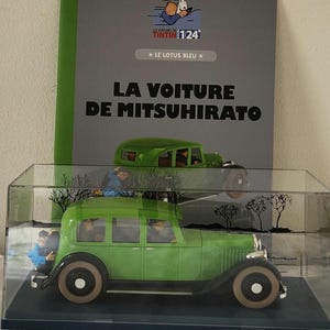 May include: A green toy car model in a clear display case, with a matching green car model on top. The display case is in front of a book cover with the text "LE LOTUS BLEU" and "LA VOITURE DE MITSUHIRATO".