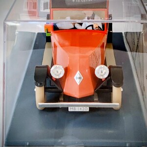 May include: A vintage-style toy car in a display case. The car is primarily orange with black fenders and cream-colored wheels. It has two headlights and a license plate that reads "MB-1420". The car is a miniature replica.