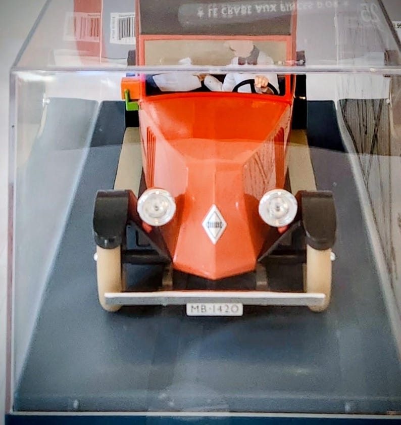 May include: A vintage-style toy car in a display case. The car is primarily orange with black fenders and cream-colored wheels. It has two headlights and a license plate that reads "MB-1420". The car is a miniature replica.