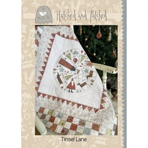 May include: A handmade quilt with a festive Christmas theme. The quilt features a central circular design with holiday figures and text, surrounded by a white background with red and green accents. The quilt is draped over a white chair.