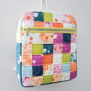 May include: A quilted Phoenix backpack pattern with a colorful patchwork design. The backpack features a yellow zipper and a white handle. The size is 16" x 12" x 4". The text on the image reads "Cotton and Joy Patterns" and "Phoenix Backpack".
